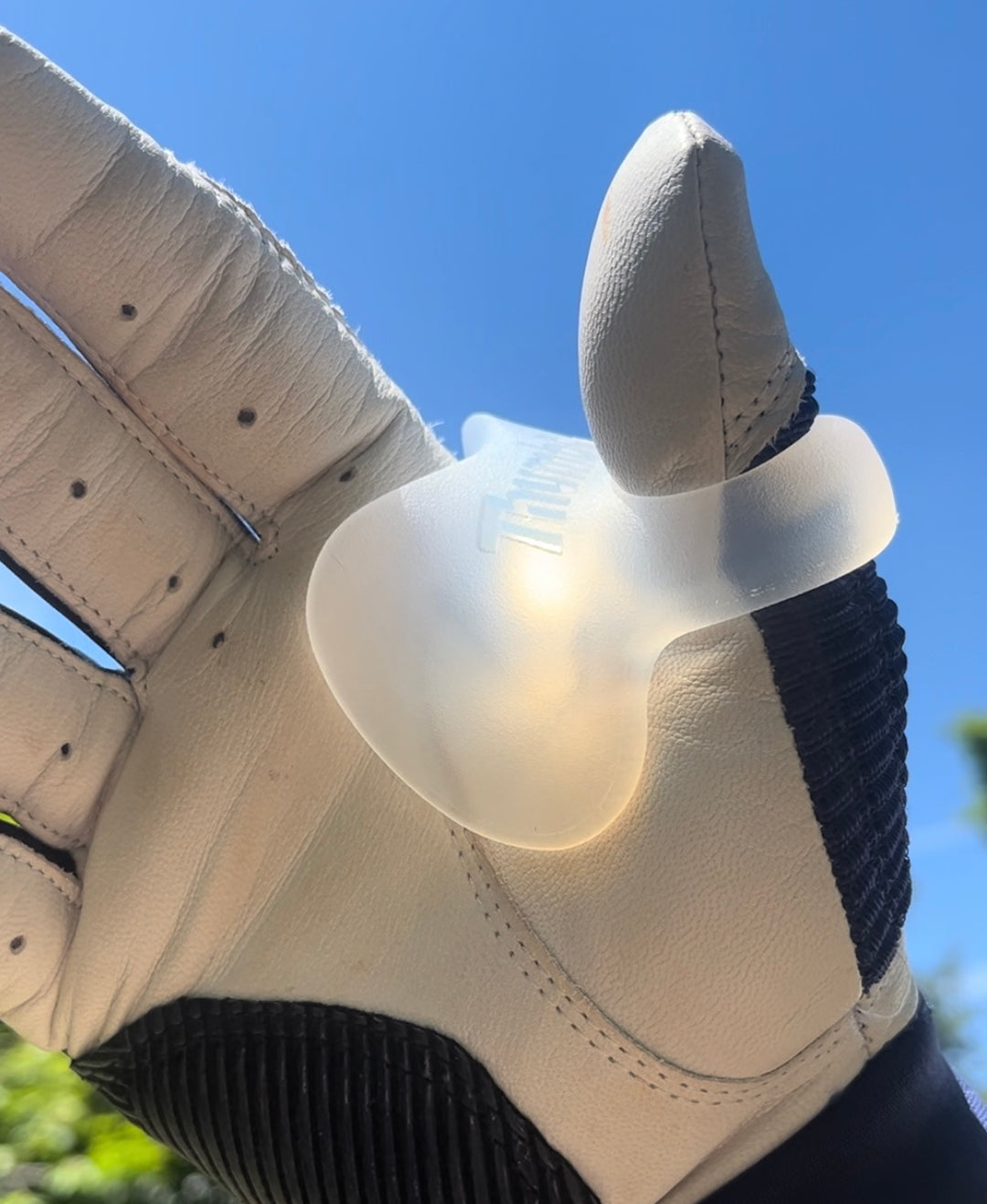 Close-up of a beige glove with a silicone baseball thumb guard against a blue sky.